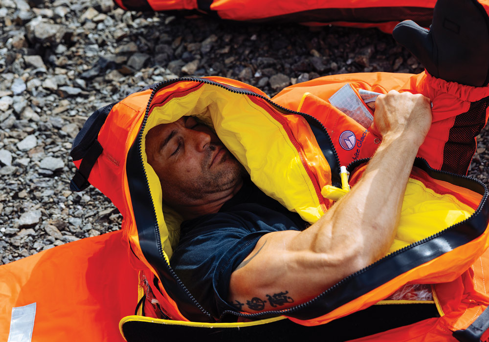 Person resting inside an orange and yellow sleeping bag on a rocky surface.
