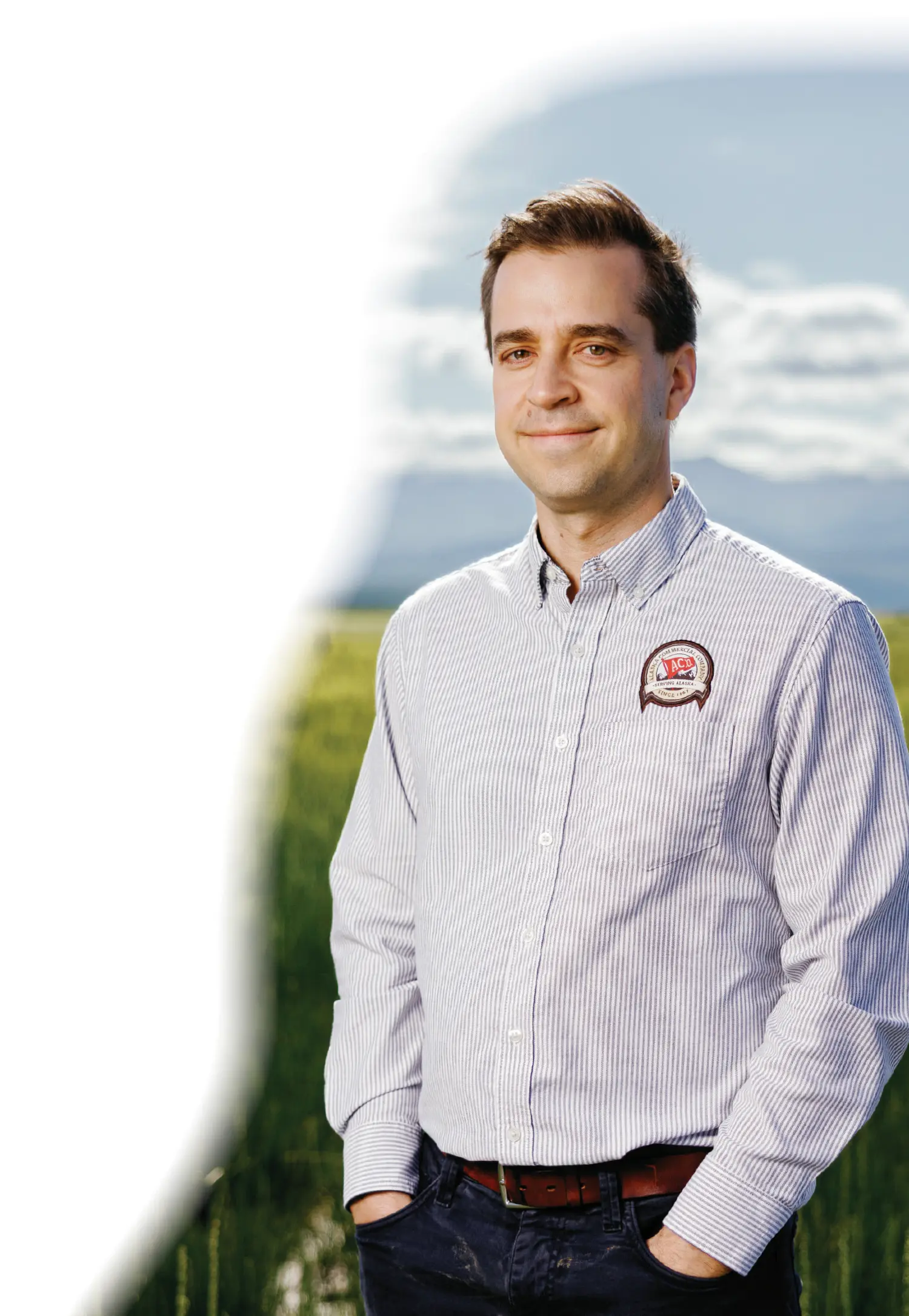 Mostly white gradient background covering the portrait orientation outdoor photograph of Kyle Hill, a smiling man who is standing amongst a grassy field and appears to have on a white button-up dress shirt that has a dark grey striped vertical orientation plaid design style pattern with an Alaska Commercial Company logo patch on the button-up dress shirt that shows the words SERVING ALASKA SINCE 1867; He is wearing dark navy blue denim jeans with his hands resting inside his pockets as he is looking towards the direction of the camera and in the blurred background behind him, mountains are visible under a blue sky with some scattered clouds on a sunny day