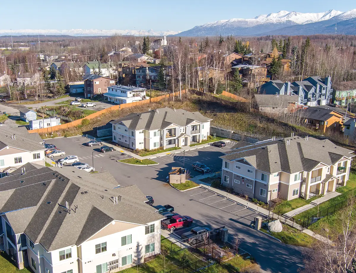 An aerial shot of new residential apartment buildings in a suburban setting, likely in Alaska. In the foreground, four apartment buildings with sloped, gray roofs and light-colored siding are visible, with cars parked in their lots. The background shows more residential homes scattered among trees, with snow-capped mountains under a clear sky in the far distance.