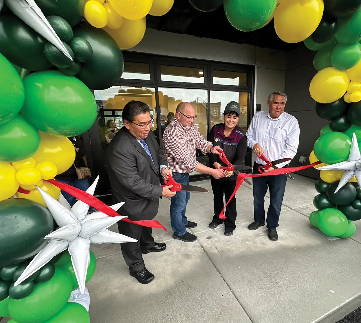Four people are shown participating in a ribbon-cutting ceremony. They are standing in front of a building entrance, each holding a pair of large scissors to cut a red ribbon. A large archway of green, yellow, and silver balloons frames the scene, with one large silver star balloon in the foreground.