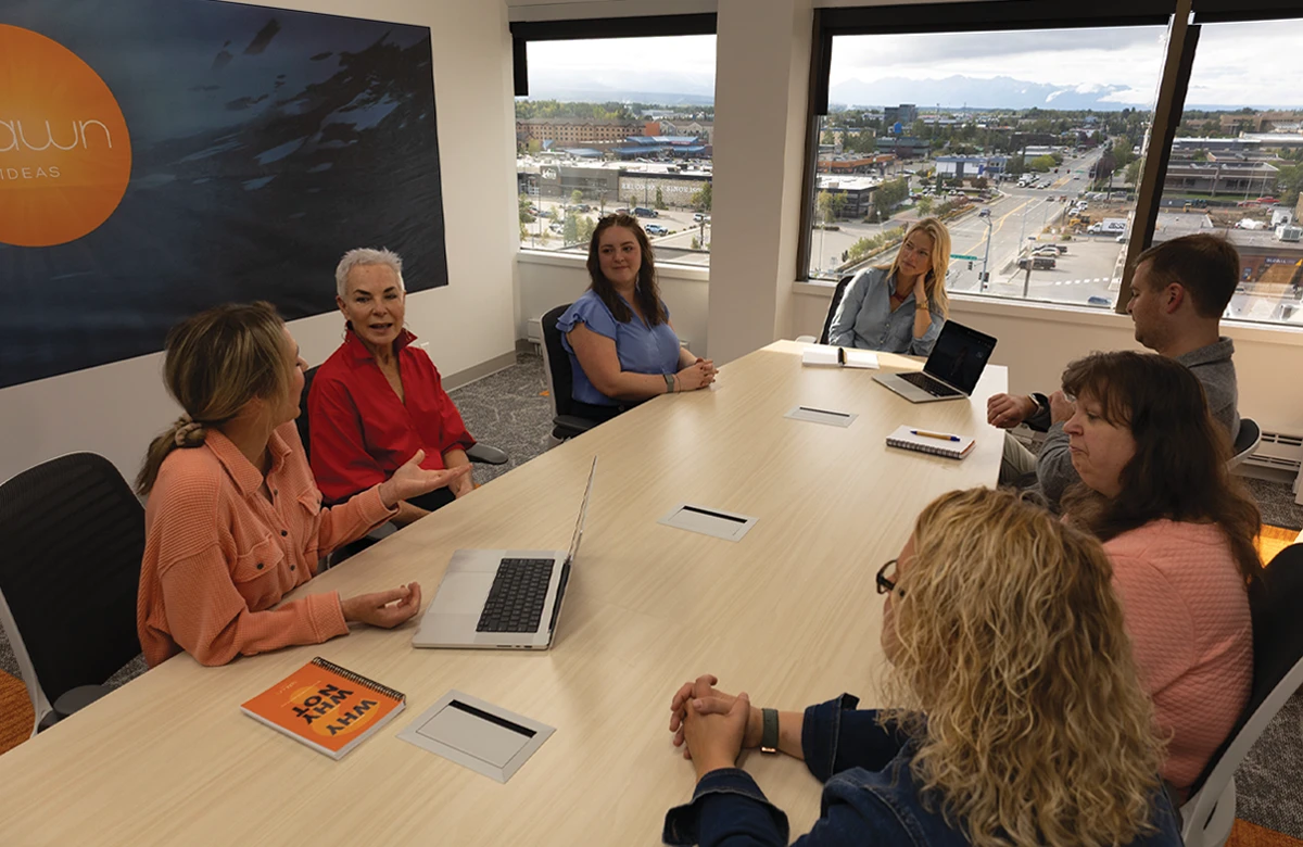 A group of seven people are sitting around a long conference table in a modern office. Some are engaged in conversation, while others are looking at laptops. The room has large windows with a view of a city street, buildings, and mountains in the distance. A logo for "Spawn" is visible on the wall.