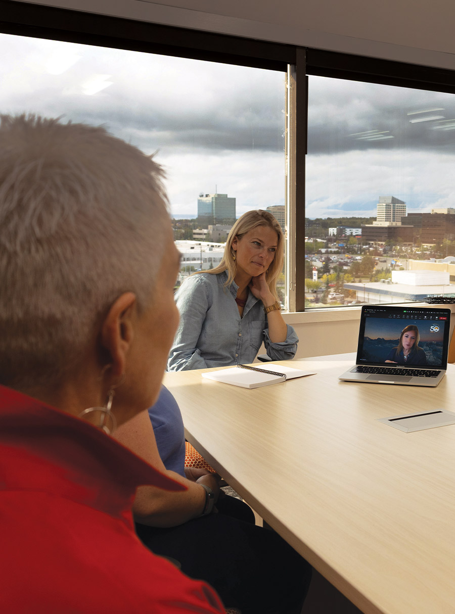 A group of three people sitting at a conference table. One person is shown from the back, another looks at a laptop on the table, and a woman on the laptop screen appears to be participating in a virtual meeting. Large windows in the background provide a view of a city skyline under a cloudy sky.