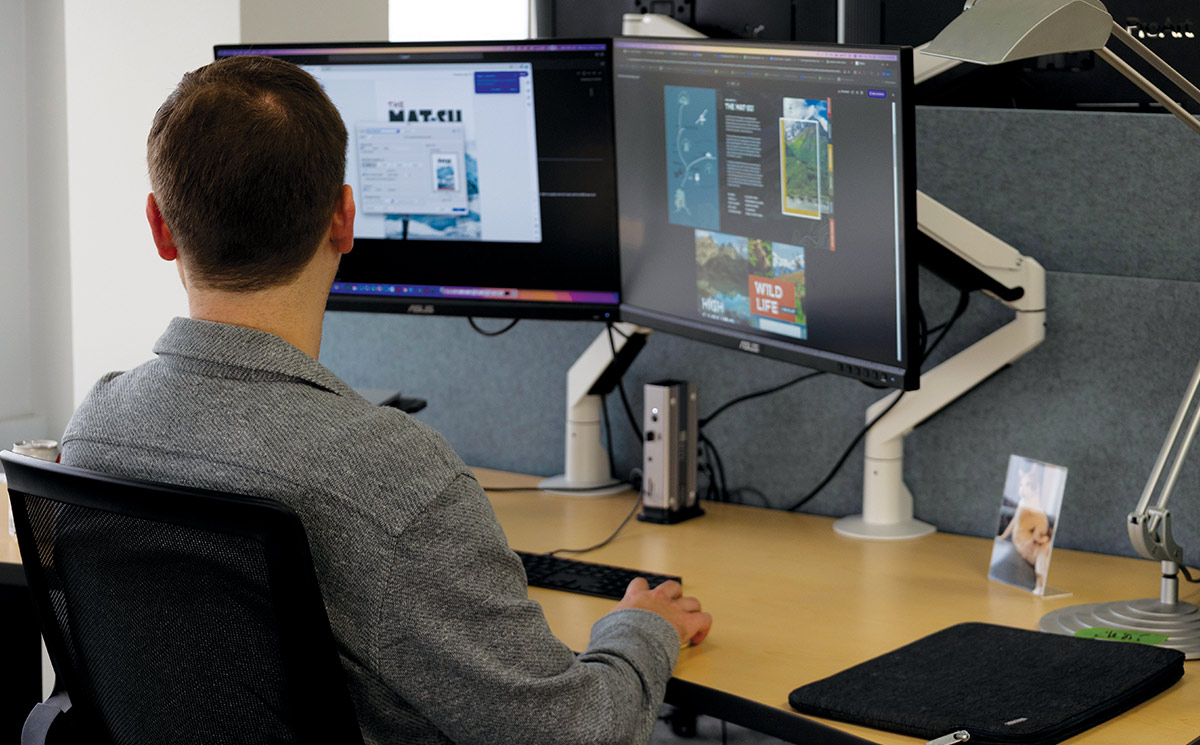 A man with his back to the camera sits at a desk, looking at two computer monitors. The screens show design work, with one displaying a website and the other showing a layout for "Wild Life." A small framed photo of a dog is on the desk to the right.