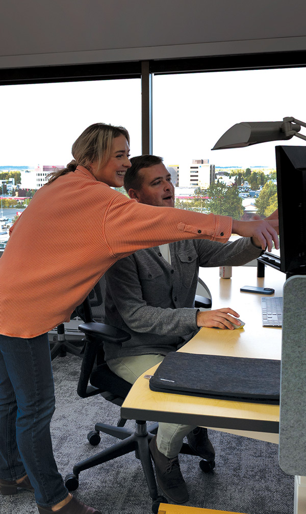A man and a woman are looking at a computer monitor at a desk. The woman, standing behind the man in a rolling office chair, is pointing at the screen, and both are smiling and engaged in discussion. The office has large windows with a view of a city landscape.