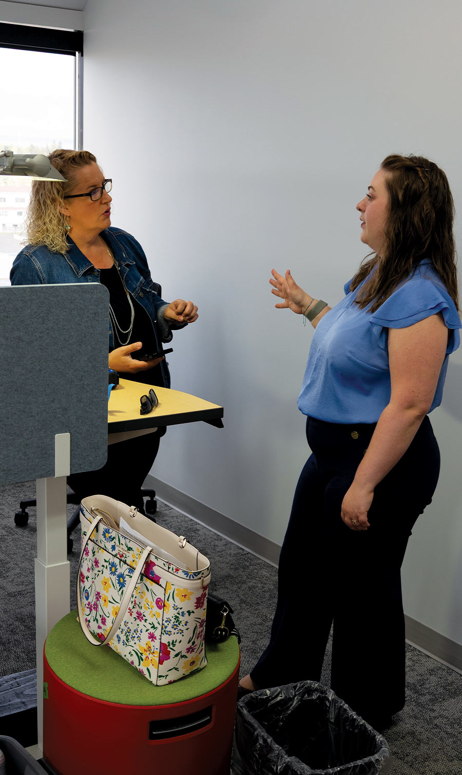 Two women are standing and talking to each other in an office setting. One woman, with curly blonde hair and wearing a denim jacket, is leaning against a desk, while the other woman gestures with her hands as she speaks. A floral patterned tote bag sits on a colorful stool in the foreground.