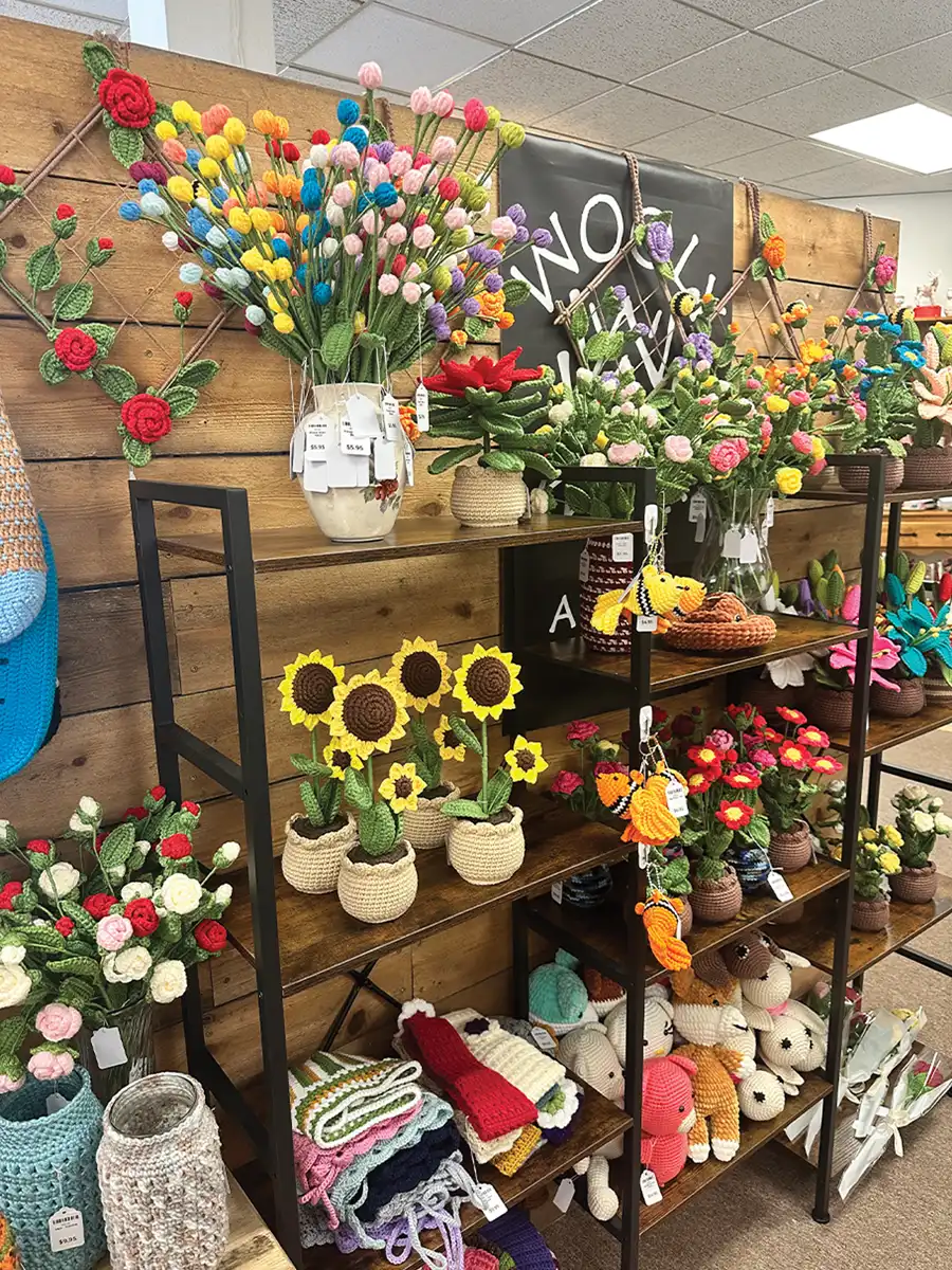 A retail display shelf filled with a variety of handcrafted yarn or crocheted items. The shelves hold an assortment of colorful flowers, sunflowers in small pots, and small stuffed animals, all made from yarn. The background wall is made of wood paneling with some crocheted decorations hanging on it.