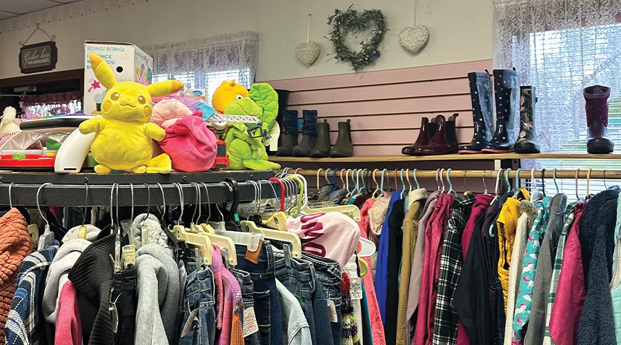 A cluttered retail display inside a store, showing racks of clothing in the foreground. Above the clothes, a shelf holds various items for sale, including plush toys like Pikachu, and several pairs of boots. Two heart-shaped decorations hang on the pink-striped wall in the background.