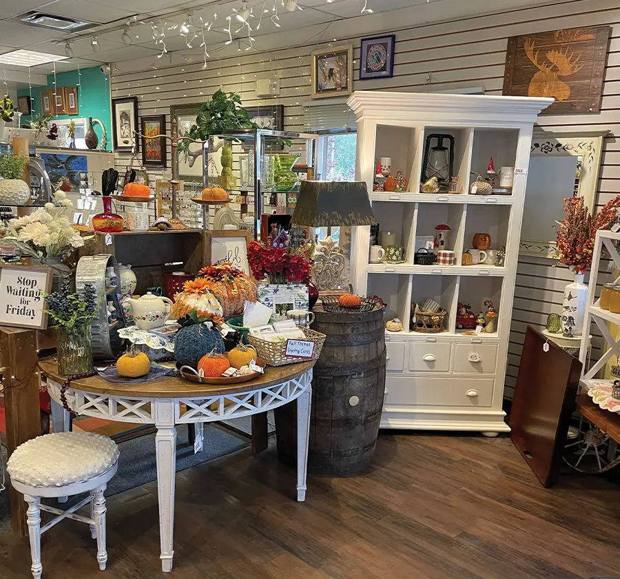 An interior view of a gift shop or boutique, with a variety of home decor and seasonal items on display. A round table in the center is decorated with artificial pumpkins and fall-themed arrangements. The room is filled with shelves and furniture holding various trinkets, lamps, and wall art, with string lights hanging from the ceiling.
