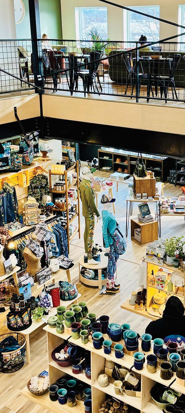 An overhead view of the interior of a two-story retail store. The ground floor is filled with a variety of goods on display, including clothing, mugs, and home goods, arranged on tables and shelves. A person with their back to the camera is browsing an item on a display stand. On the second floor, a cafe seating area with tables and chairs is visible.