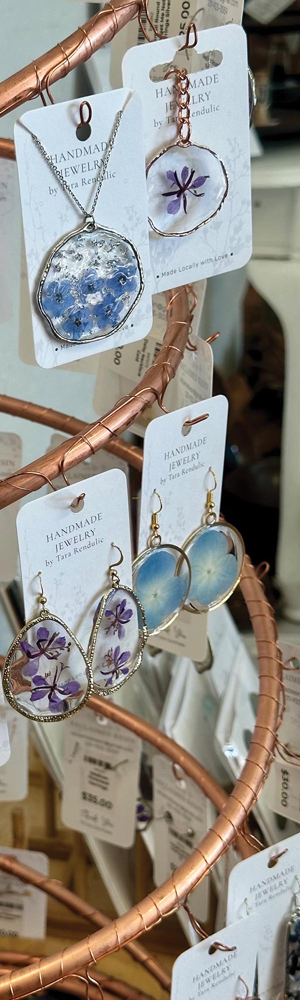 A close-up of a copper-colored jewelry stand, holding several pairs of earrings and necklaces. Each piece of jewelry features real pressed flowers, such as blue and purple forget-me-nots, encased in a clear resin and metal frame.