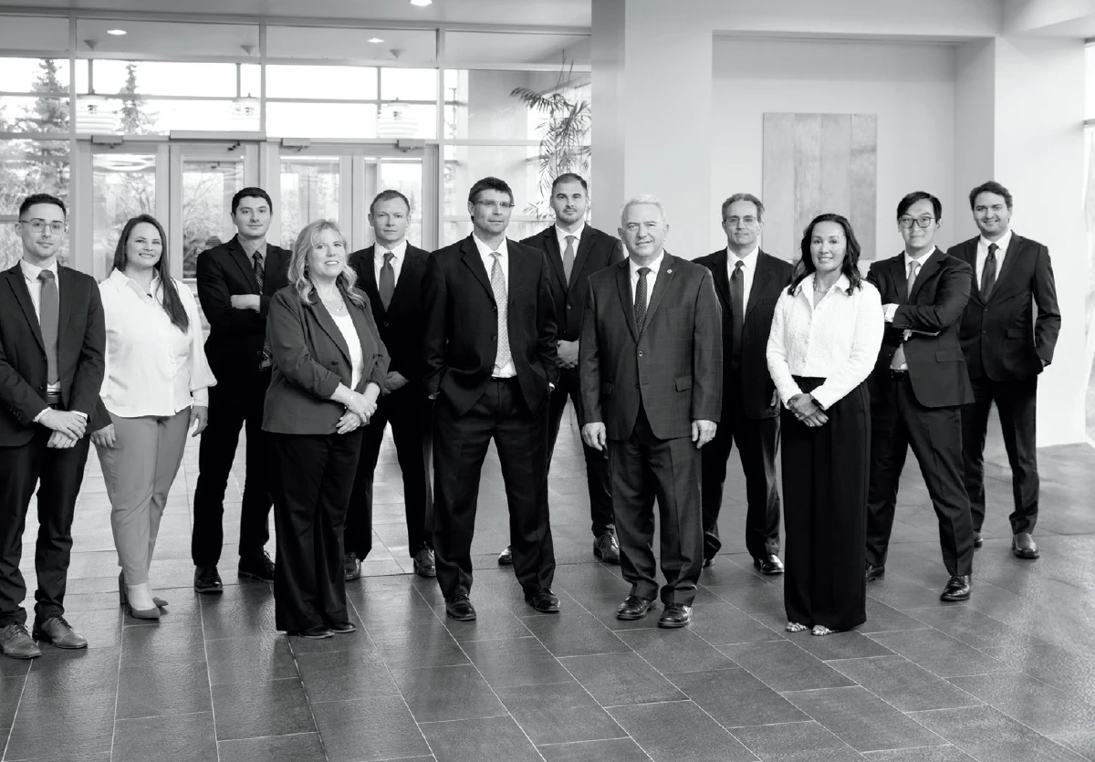 A black and white photo shows a group of 11 professional people, dressed in business attire, standing in a modern office lobby. The group includes both men and women, who are smiling and posing for the photo. 