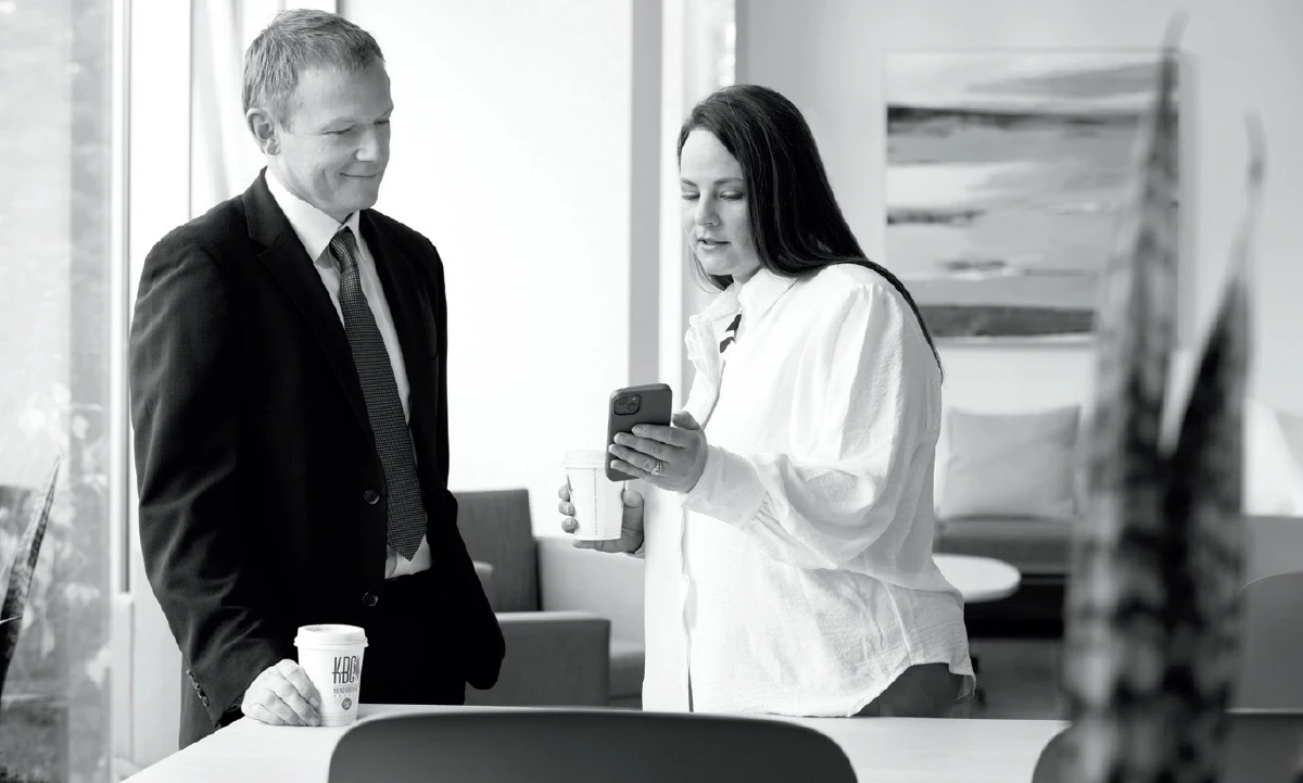 A black and white photo shows a man and a woman in an office setting. The man, wearing a suit and tie, is looking at the woman who is holding a coffee cup and looking at her phone. They appear to be in a casual conversation, possibly on a break.