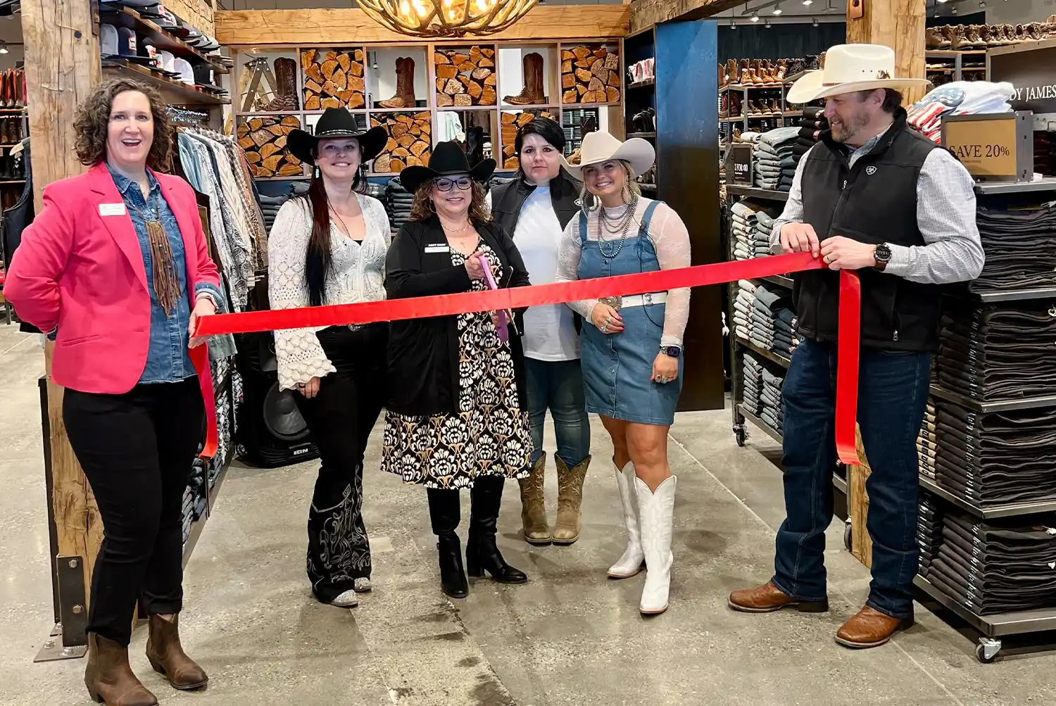 A group of five women and one man stand in a western-style retail store, holding a red ribbon for a ribbon-cutting ceremony. The people are smiling, and most are wearing cowboy hats and boots. The store shelves in the background are filled with clothing and boots.