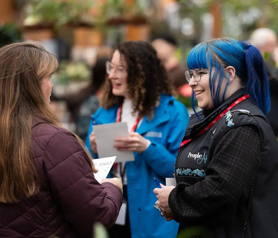A group of three people is engaged in conversation at a professional networking event. Two women, one with long brown hair and another with bright blue hair, are smiling at each other while the third person, in a blue jacket, stands in the background.