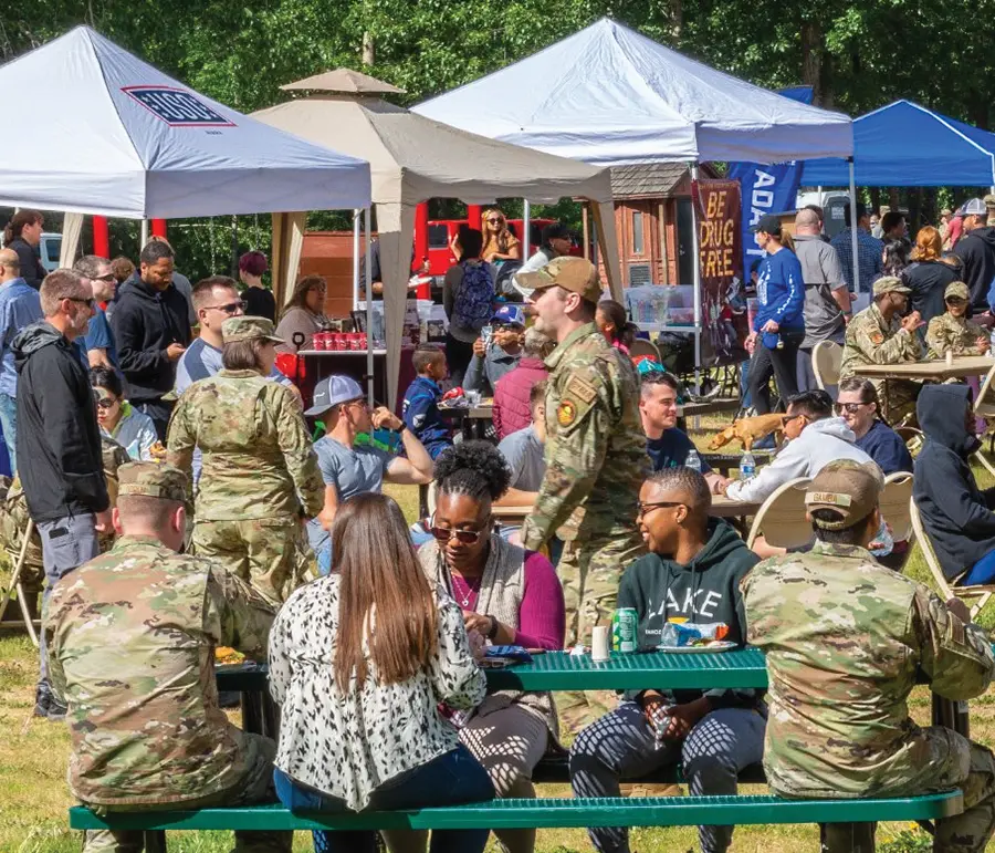 A wide shot shows a lively outdoor event with many people gathered under white tents. Some attendees are in military fatigues, while others are in casual clothes. People are sitting at green picnic tables, walking around, and mingling. Banners and various booths are visible in the background, suggesting a community fair or gathering.