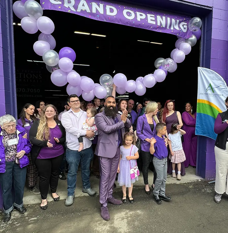 A diverse group of people, many wearing different shades of purple, are gathered outside a building entrance decorated with a garland of purple and silver balloons. A banner above them reads "GRAND OPENING." An Anchorage Chamber flag is visible to the right. A man in a purple suit is clapping, while a person in the foreground holds a baby.