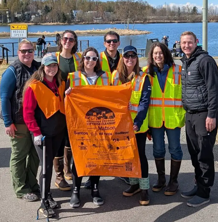 A group of eight volunteers, wearing bright yellow or orange safety vests, stand together for a photo next to a body of water on a sunny day. They are holding a large orange trash bag that is covered with logos from sponsors like Santos, Matson, and Fred Meyer. The group appears to be participating in a community cleanup event.