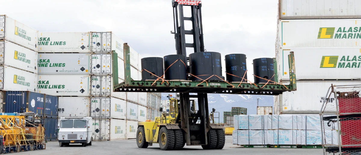  A large yellow forklift is lifting a flatbed trailer loaded with several large, cylindrical black objects, likely industrial supplies, at a shipping yard. In the background, stacks of white Alaska Marine Lines shipping containers and a smaller white vehicle are visible under a cloudy sky.