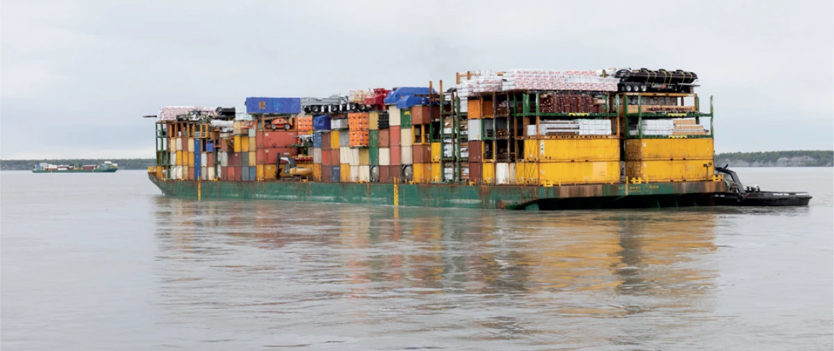  A long barge is heavily loaded with stacks of multicolored shipping containers and other cargo, being pushed by a small tugboat at its stern. The barge is floating on a wide river under a gray sky. Another, smaller barge is visible in the distance on the left.