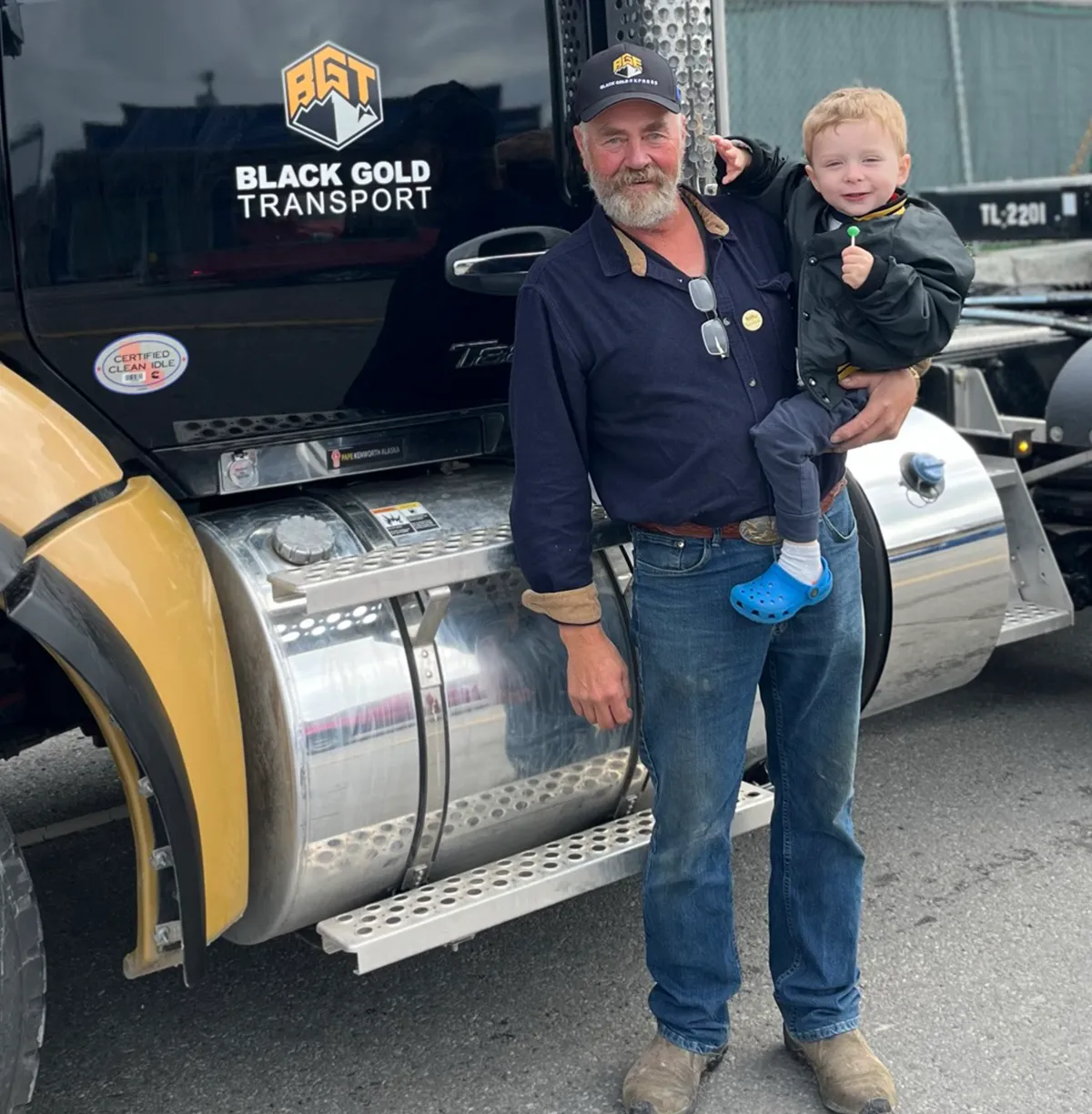 Jim Huffman man in a baseball cap, dark blue shirt, and jeans holding his son in his arms. He a full beard and is standing in front of a large black and gold Black Gold Transport semi-truck. The boy is wearing a black jacket and blue Crocs. They both appear to be happy and are posing for the photo.