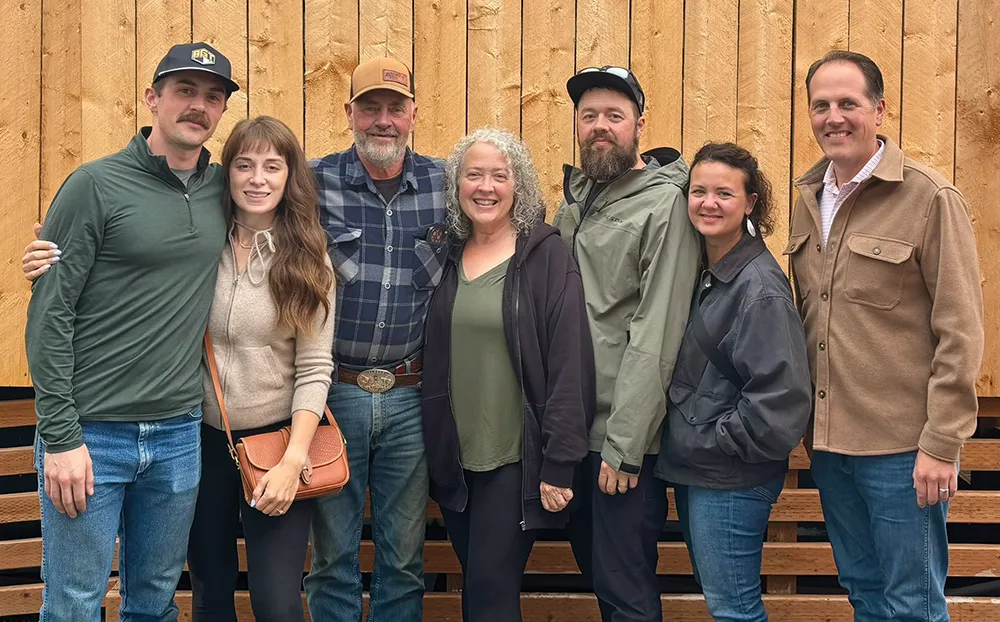 A family of seven, consisting of two older parents and five adult children, is standing side by side in a casual group photo. They are all smiling and posing in front of a rustic, wooden-planked wall. The individuals are wearing a mix of casual clothing, including jeans, hoodies, and button-up shirts.