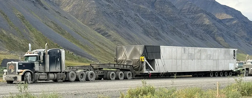 A long-haul semi-truck, with a black and gold Black Gold Transport logo on its door, is pulling a massive piece of equipment on a flatbed trailer. The equipment is partially covered by a tarp. The truck is driving on a dirt road through a mountainous landscape in Alaska, with green and brown hills visible in the background.