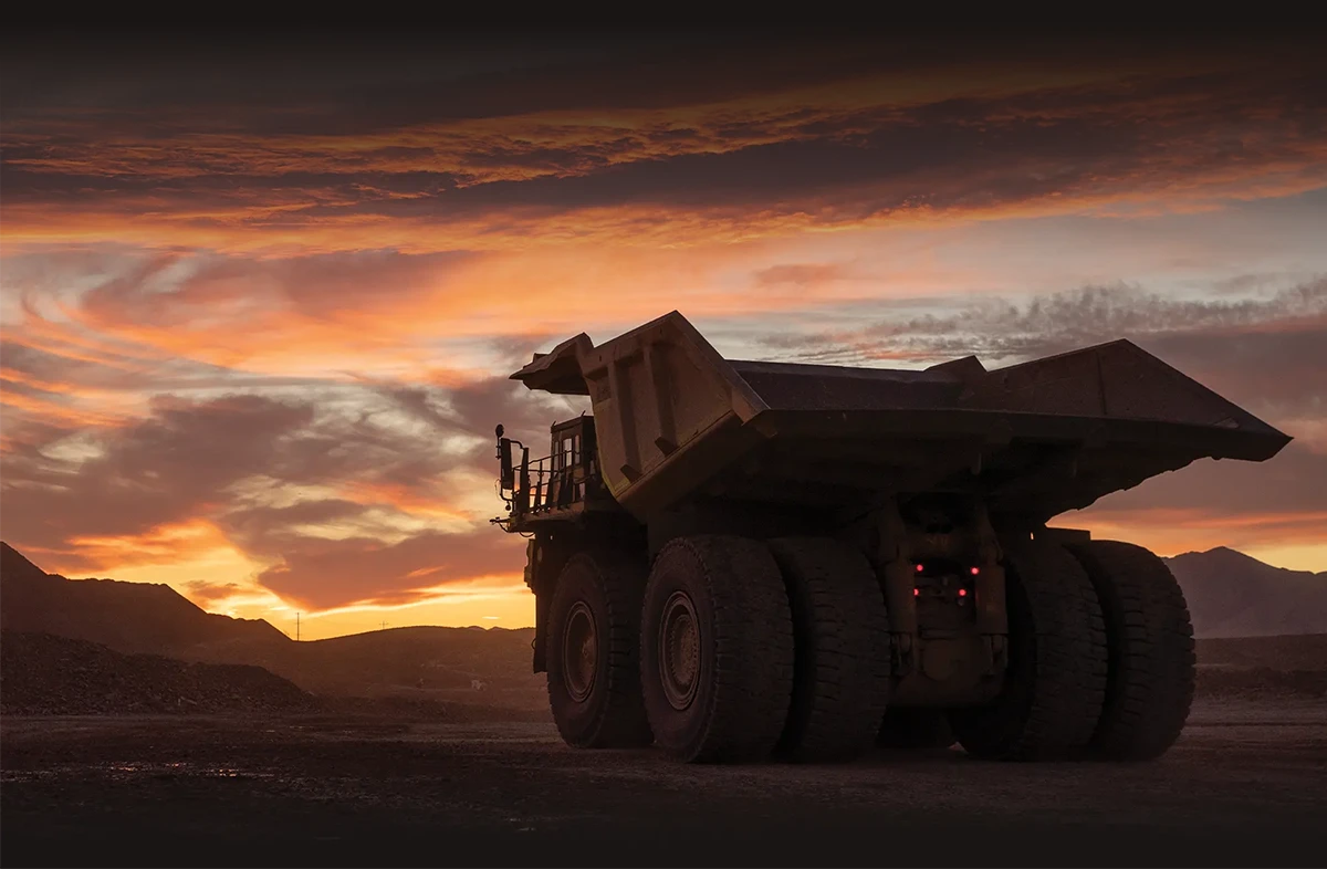 A large mining dump truck is silhouetted against a dramatic sunset with orange and red clouds. The truck is positioned on a dirt road at a mine site, and its rear brake lights are on. The horizon features mountains and hills, adding to the rugged, industrial landscape.