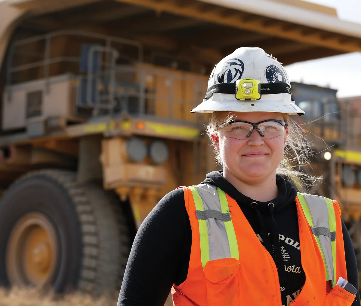 Judy Patrick wearing a white hard hat with a headlamp, safety glasses, and a bright orange safety vest over a black hoodie. She is standing in front of two large yellow dump trucks.