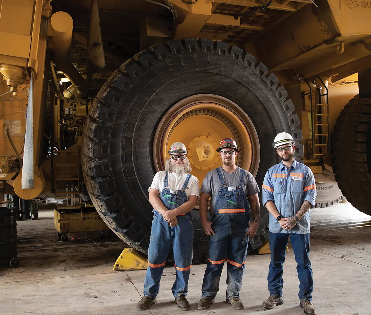 Three employees, wearing hard hats, safety glasses, and denim overalls, stand in front of a giant truck's tire. Two of the men have beards, and all three are looking at the camera. They appear to be in a garage or maintenance facility.