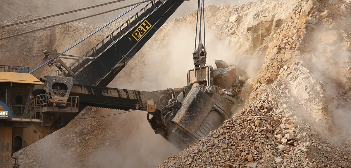 A massive P&H mining shovel with a large bucket scoops up rock and dirt from the side of a quarry. The scene is dusty with debris flying around the machine as it operates. The shovel is black with yellow lettering, contrasting with the light brown and tan rocks and dirt of the mine.