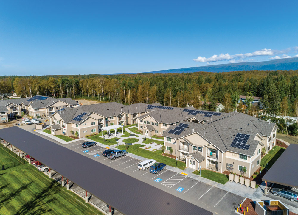 A high-angle view of a new, two-story apartment complex with a central courtyard and covered parking. Solar panels are visible on many of the rooftops. The buildings are tan with dark gray roofs.