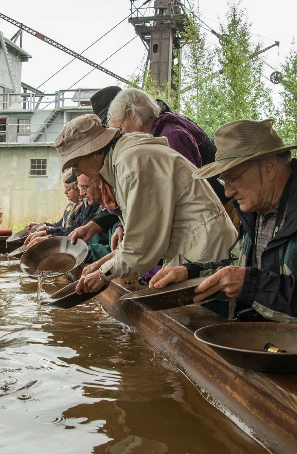 People panning for gold at a water trough with industrial machinery in the background.