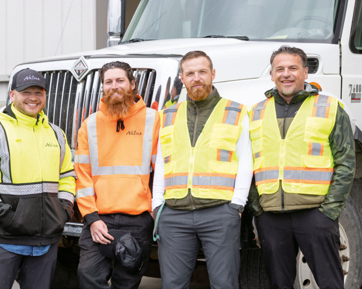 The image features four men standing side-by-side in front of a white truck. They are all wearing construction or work gear, including high-visibility jackets and vests.