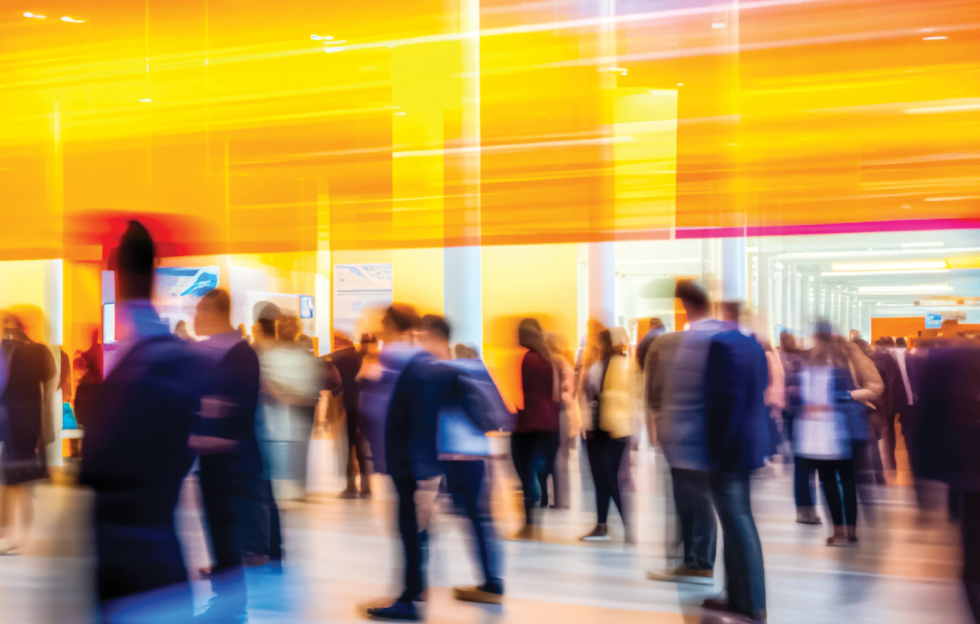 The image shows a large, crowded space, likely an event or conference, with a long-exposure effect that blurs the movement of people and lights. The people are depicted as streaks of motion, suggesting a bustling, fast-paced environment.