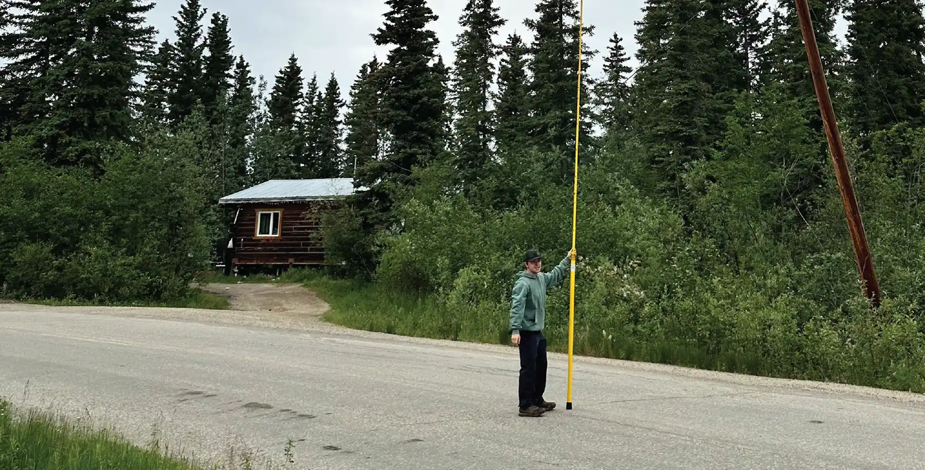 The image shows a person standing on the side of a rural road, holding a long, vertical yellow pole. They are wearing a green hooded sweatshirt and a hat. In the background, there is a small wooden cabin with a metal roof, partially obscured by lush green trees and bushes. The setting appears to be a remote, forested area.