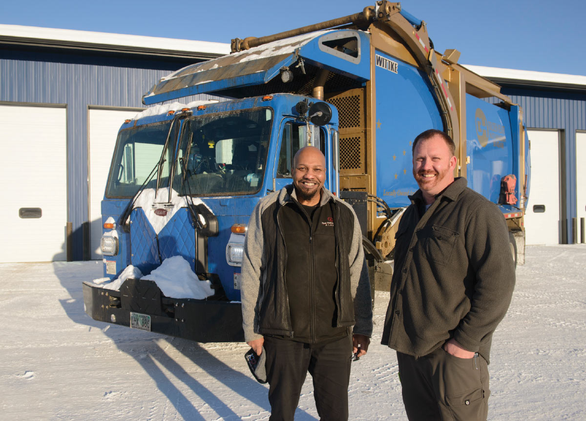 Two men are smiling and standing in front of a blue and white garbage truck. The ground is covered in snow.