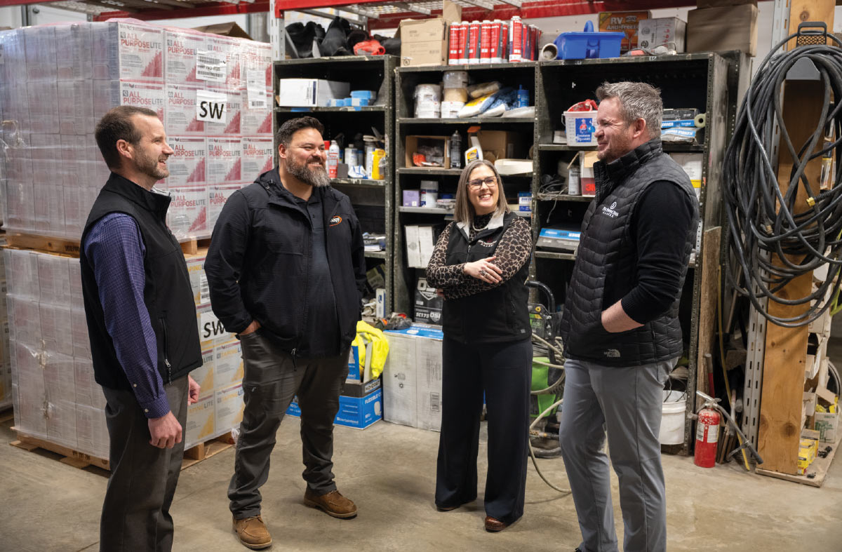 Four people are standing and talking inside a warehouse. They are surrounded by shelves and stacks of boxes.