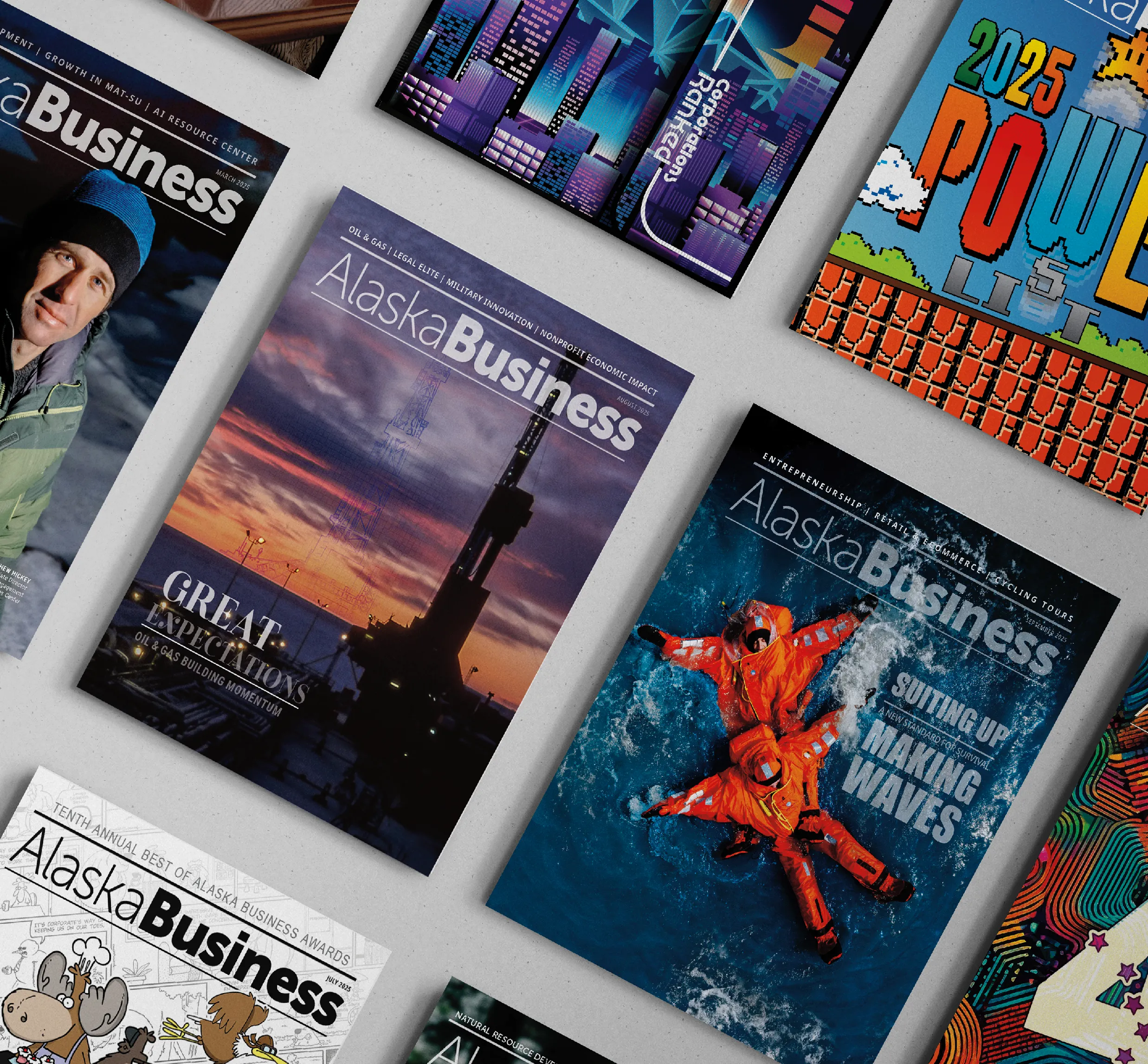 Close-up of several Alaska Business magazine covers arranged on a tabletop, including an oil rig at sunset with the headline “Great Expectations” and a rescue training cover featuring two people in bright orange immersion suits floating in water.
