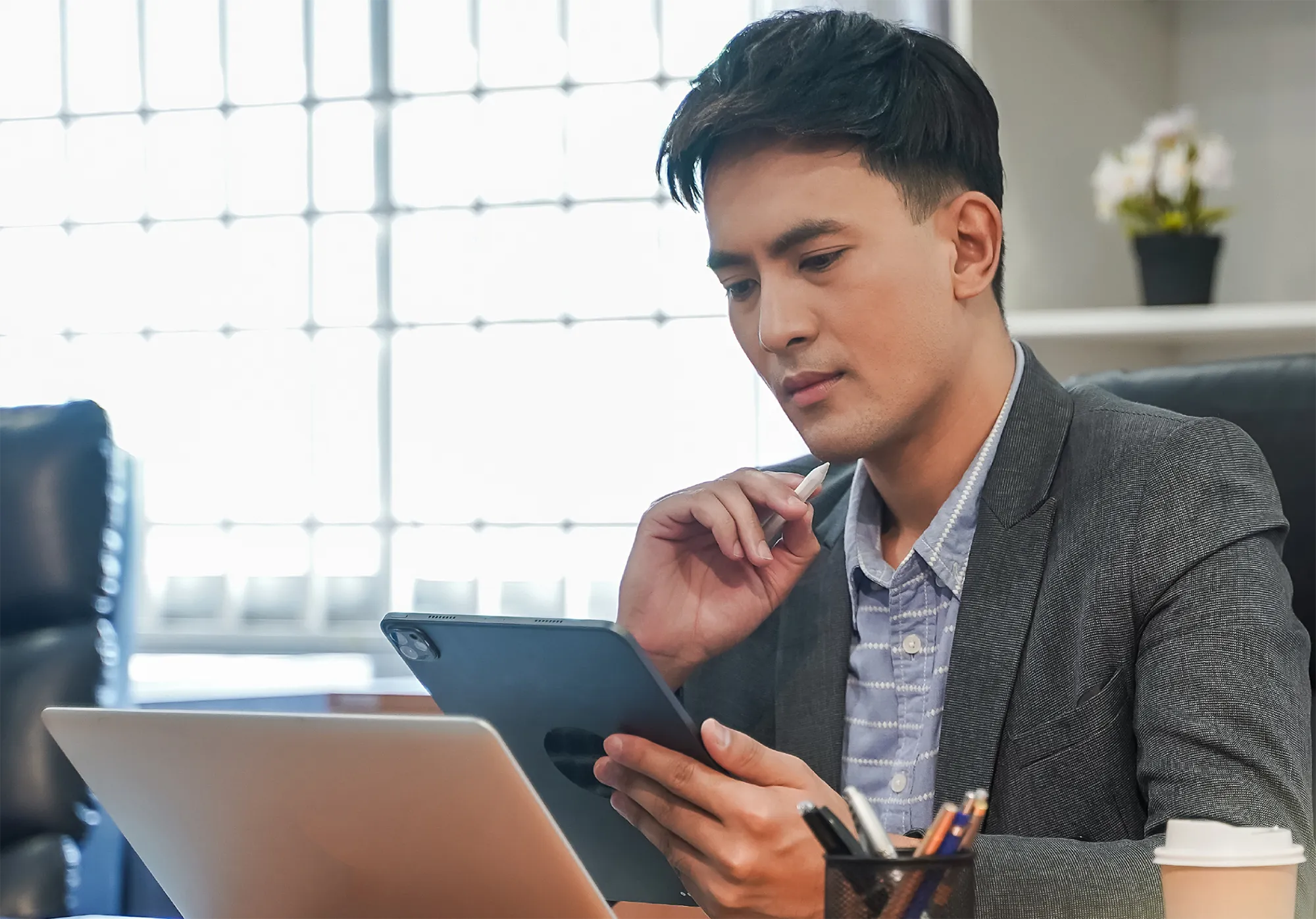 Business professional sitting at a desk with a laptop and tablet, holding a stylus and reviewing digital content in an office setting.