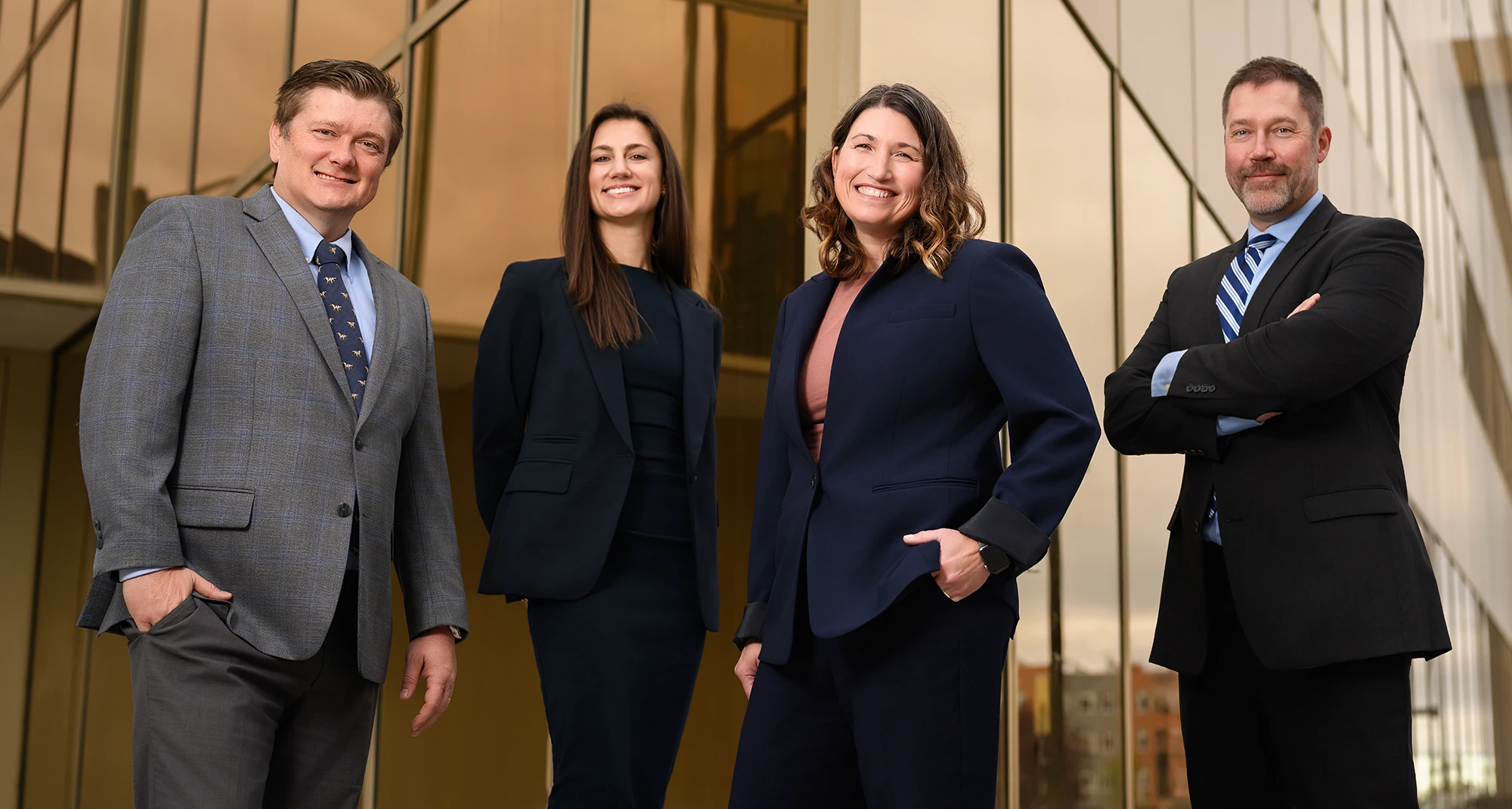 Four professionals in business attire stand smiling outside a modern glass building, posed together as a group portrait.