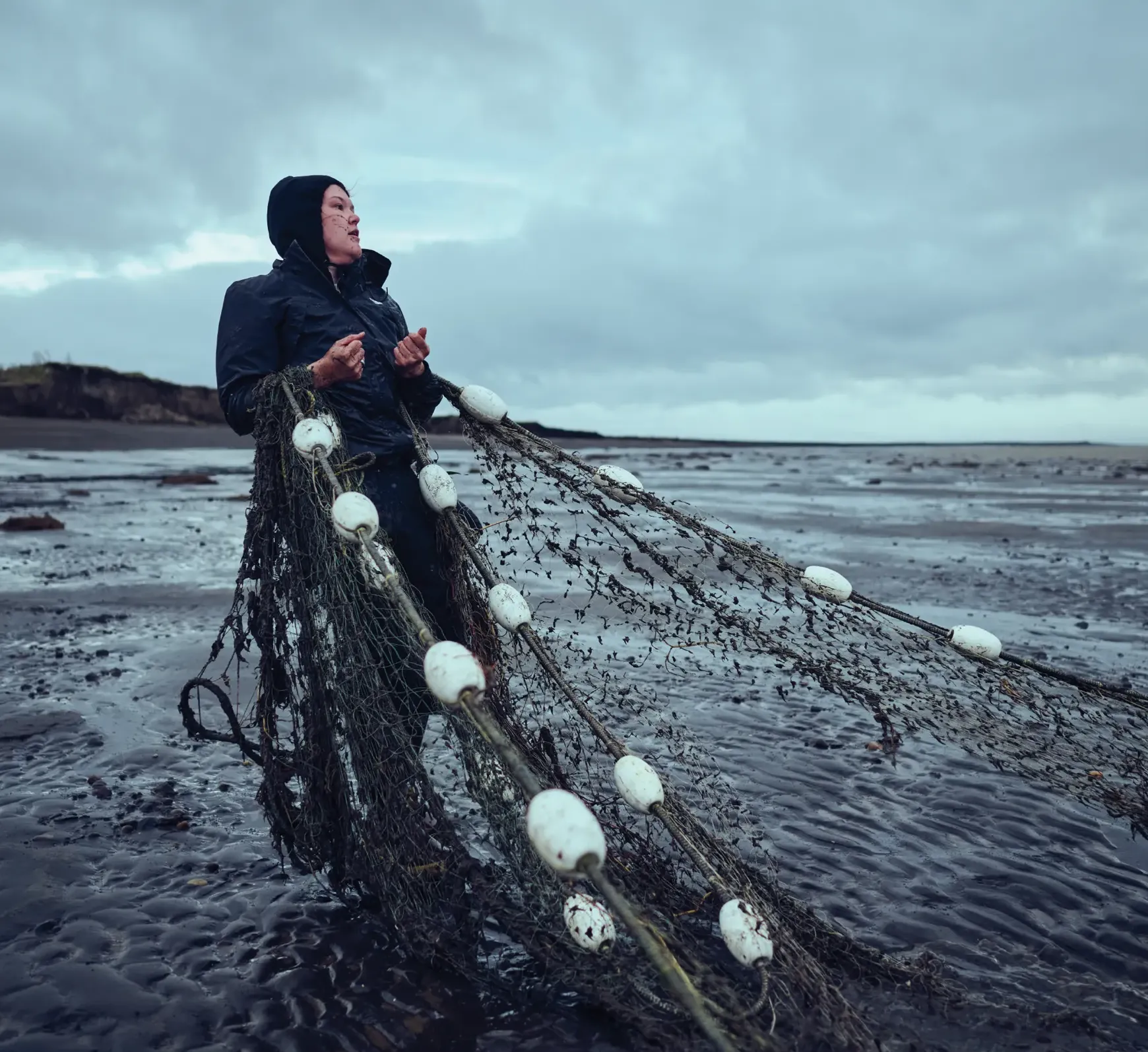 Person in a dark jacket stands on a wet shore, holding a fishing net with white floats against a cloudy sky.