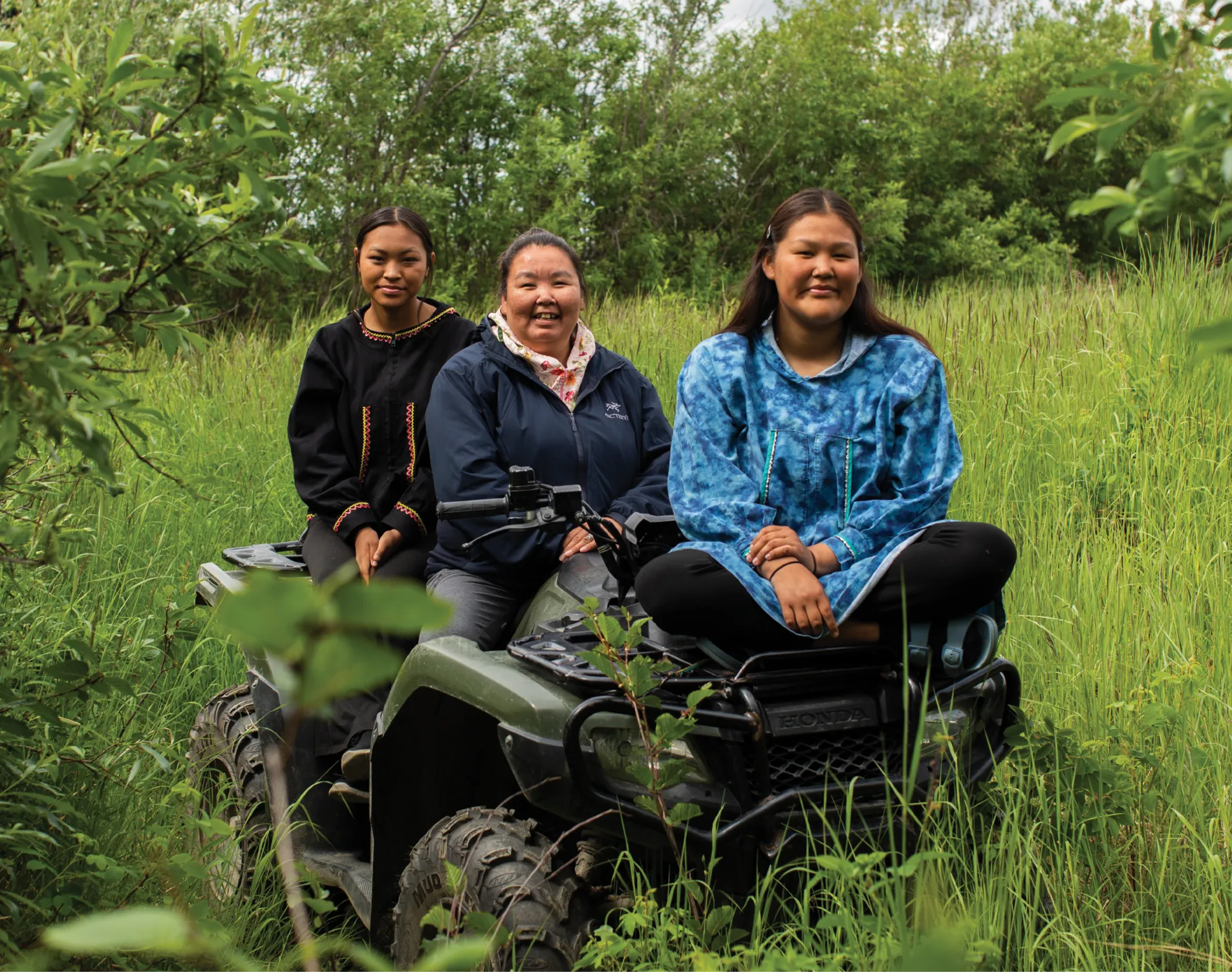 Three women sit on an ATV in a grassy area surrounded by greenery, wearing traditional clothing.