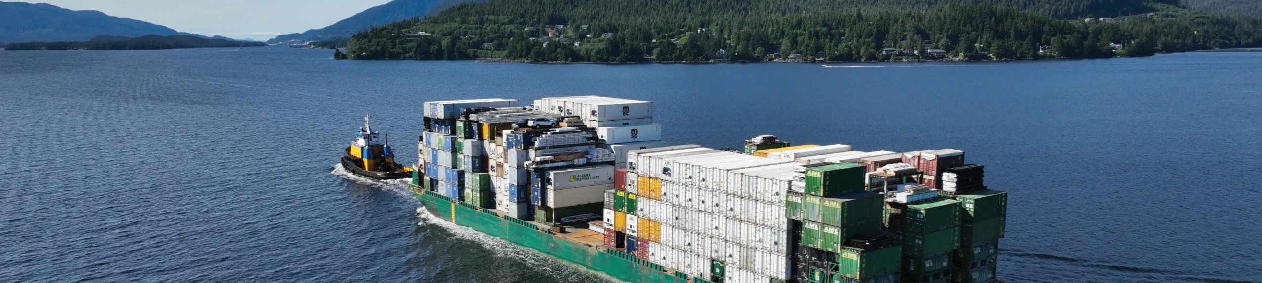 Tugboat guiding a large cargo ship loaded with colorful shipping containers through calm waters surrounded by forested hills.