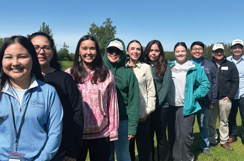 Group of diverse individuals standing outdoors in a line, smiling against a clear blue sky and green landscape.