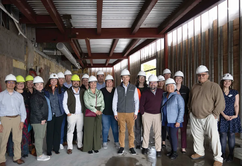 Group of people wearing hard hats posing inside a construction site with metal beams and walls in the background.