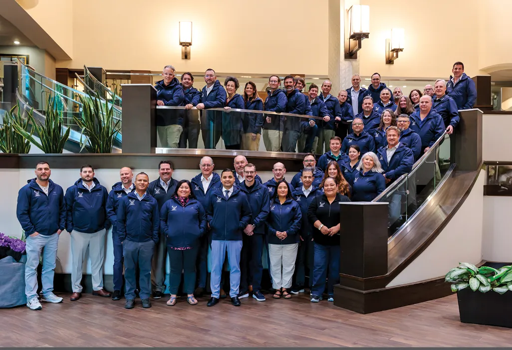 Group of people in matching navy jackets posing on a staircase in a modern building with plants in the background.