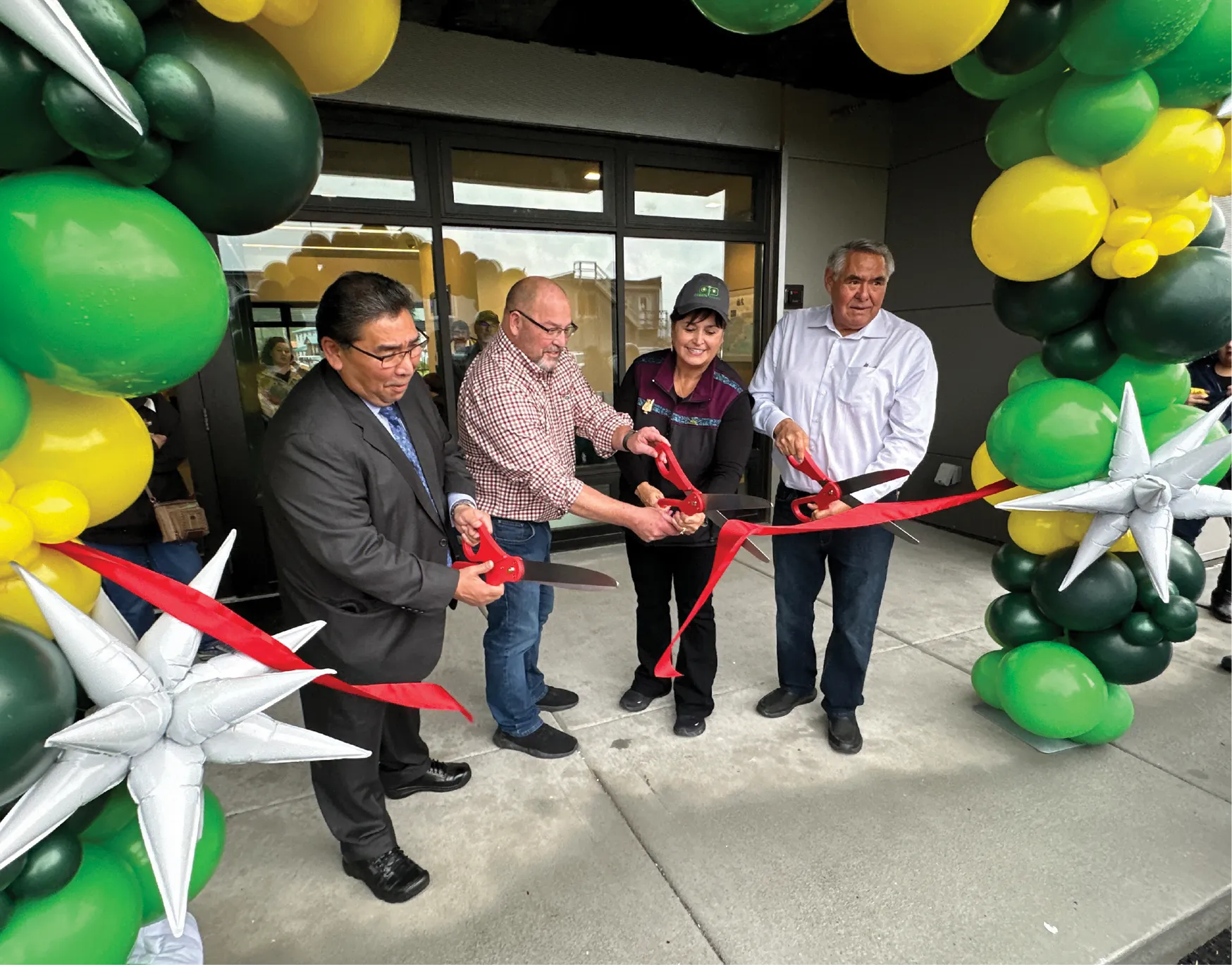 Four people cutting a red ribbon at an entrance decorated with green, yellow, and silver balloons.
