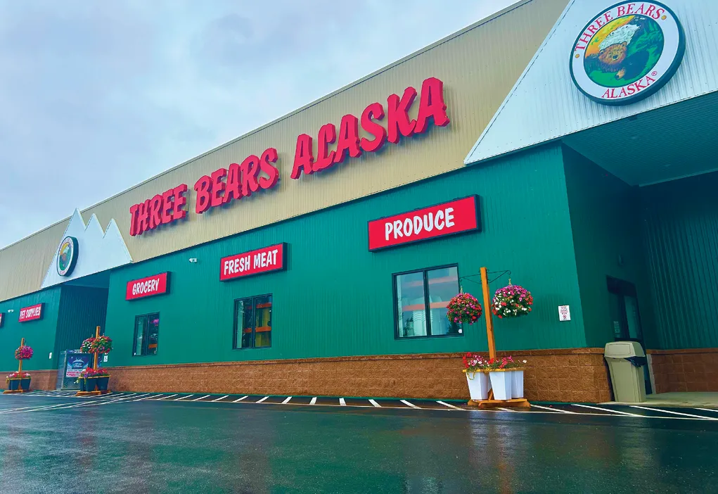 Storefront of Three Bears Alaska with signs for Grocery, Fresh Meat, and Produce, featuring hanging flower baskets.