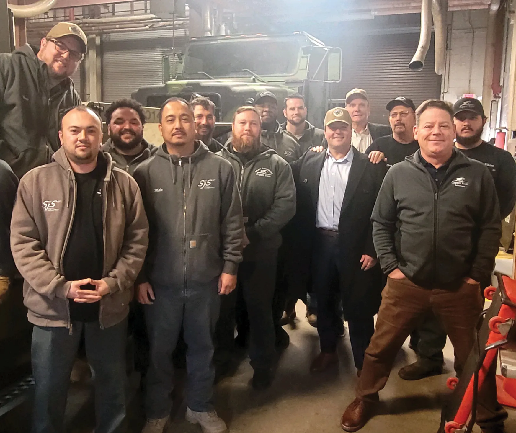 Group of people in gray jackets posing together in a workshop with military vehicle in the background.