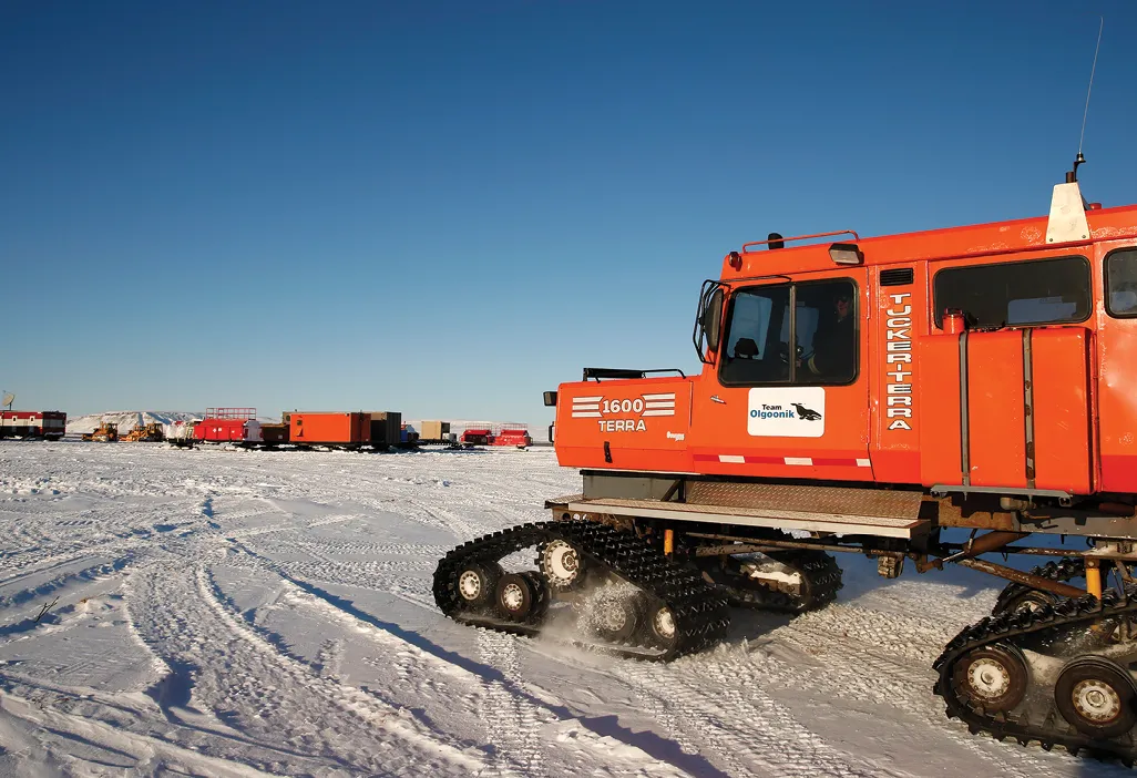 Orange tracked vehicle on snow with blue sky and distant red structures in the background.